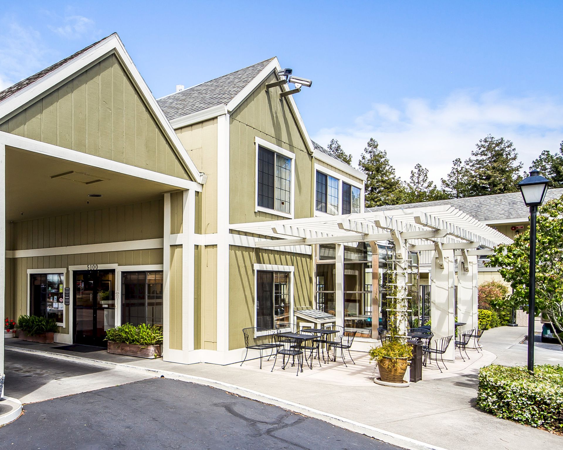 A light olive-green, two-story building with beige trim, a covered entry, outdoor seating with metal tables, and a glass atrium extending along the side.