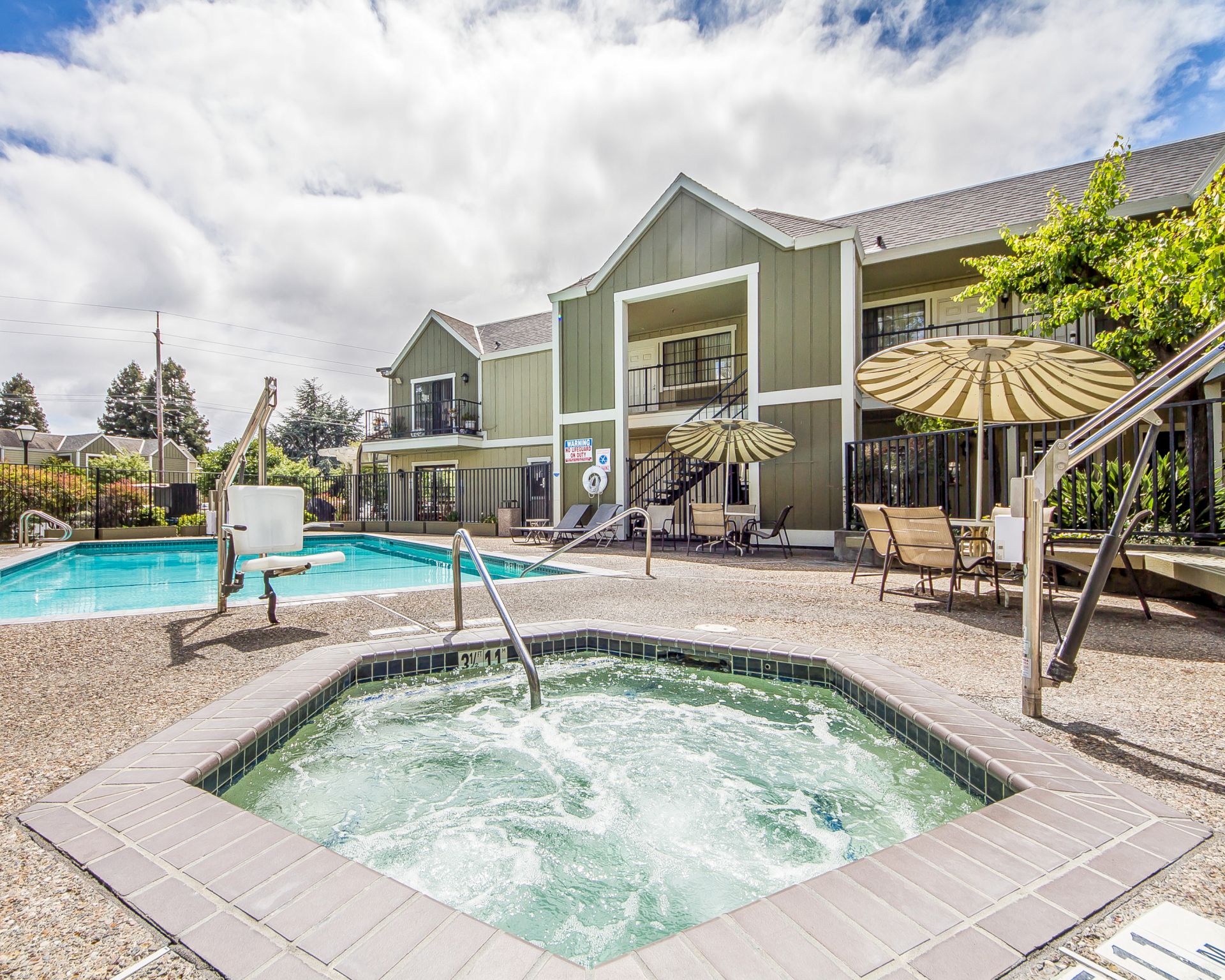 A sunny poolside retreat at a green hotel-like complex, with a hot tub in the foreground, lounge chairs, umbrellas, and a two-story building in the background.