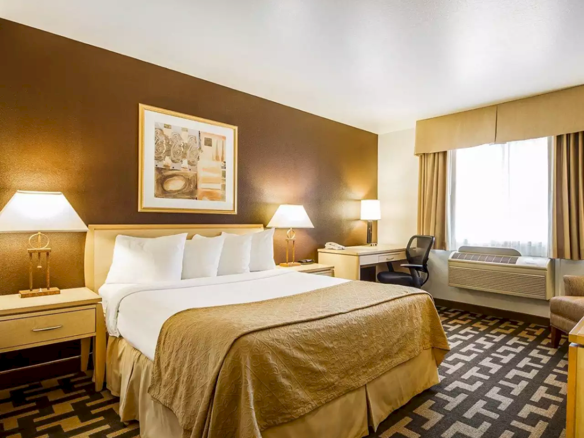 A hotel room with a made bed, bedside lamps, curtains, desk, and a window A/C unit on a patterned carpet floor.