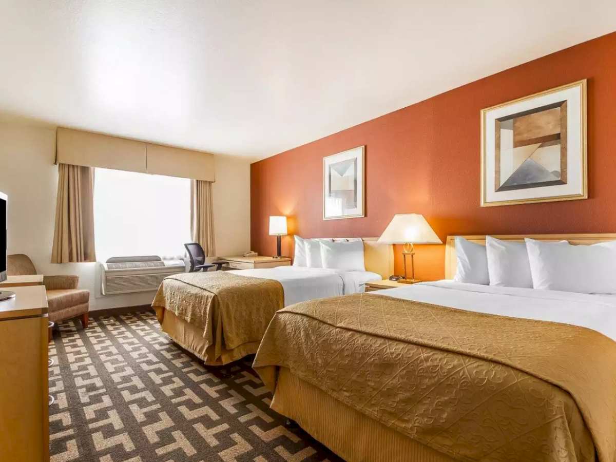 A modern hotel room with two beds, orange wall, framed art, lamps, and patterned carpet.