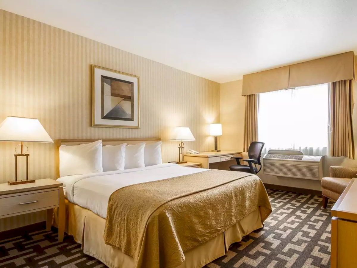A hotel room with a neatly made bed, bedside lamps, a desk chair, an air conditioner, and gold curtains by the window.