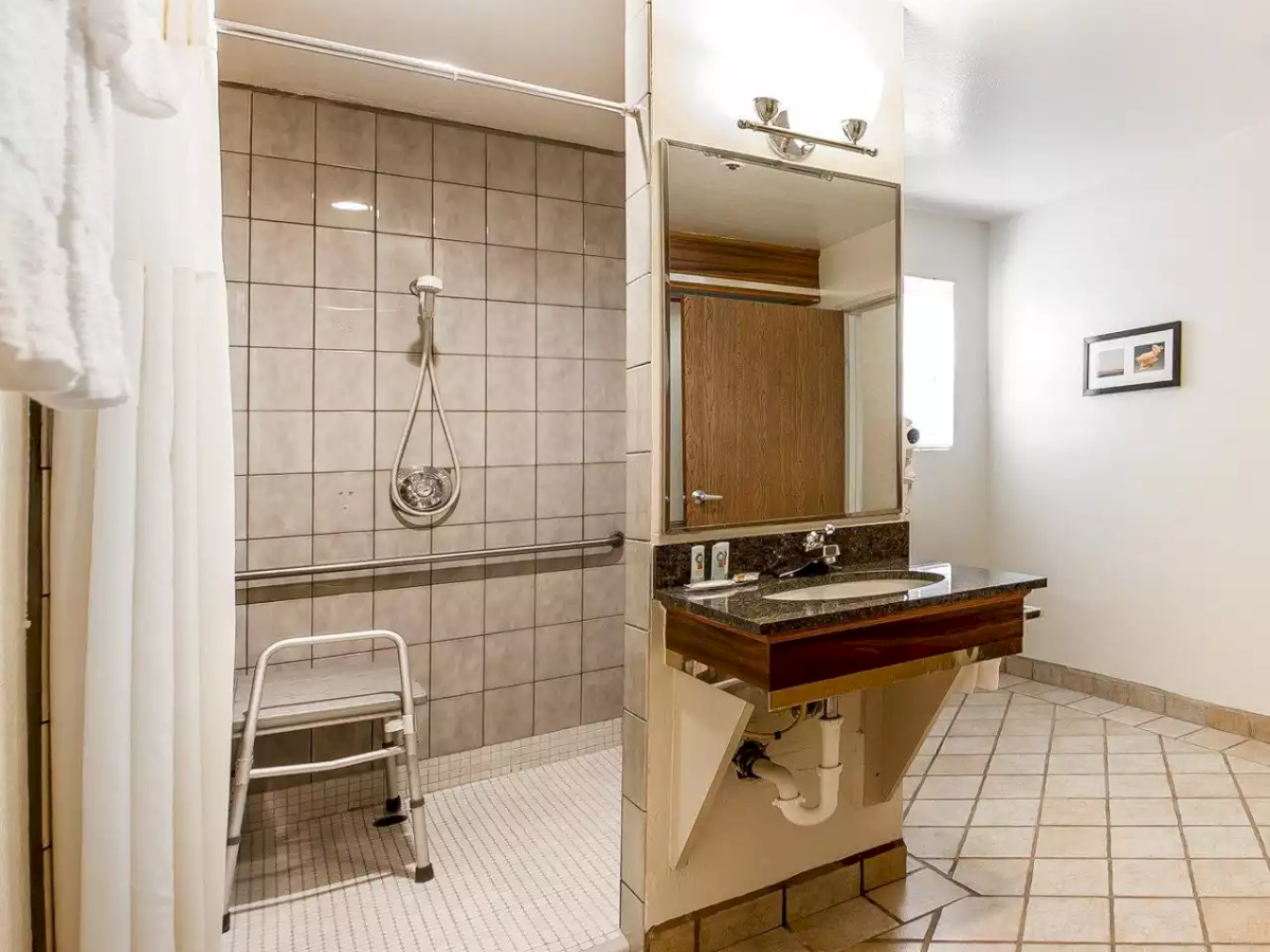 A modern accessible bathroom with a tiled shower, handheld shower rail, metal shower chair, and a granite vanity sink.