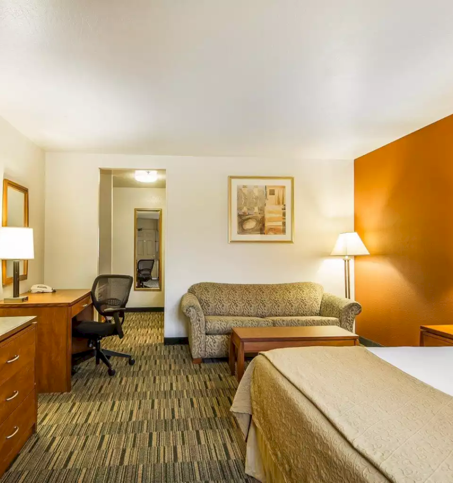 A hotel room with a bed, sofa, desk, TV, lamps, and neutral decor.