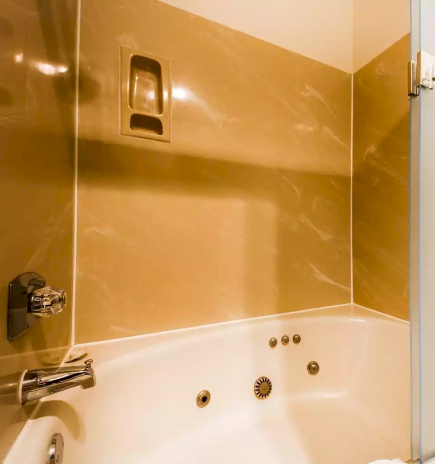 A modern bathroom corner with glossy gold tiled walls, a white bathtub, and a chrome faucet and shower panel, ending.