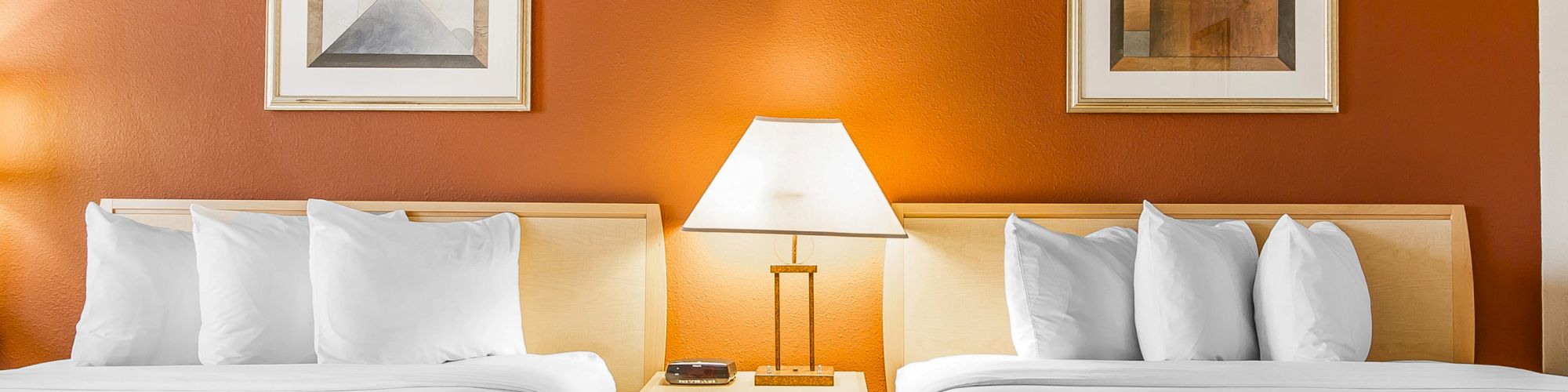A hotel room with two neatly made beds, an end table with a lamp, and framed abstract art on an orange wall.