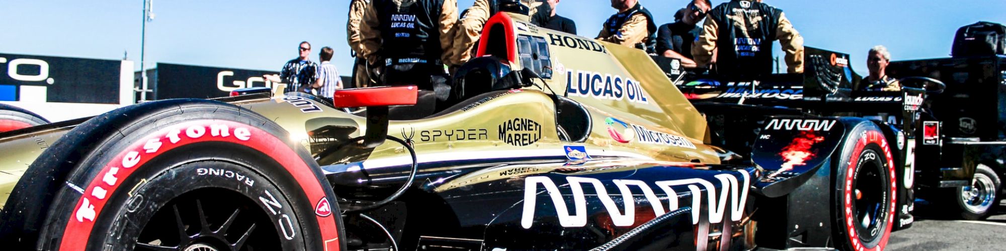 Race team members in helmets crowd around a Lucas Oil&ndash;liveried IndyCar on a track, readying it for action.