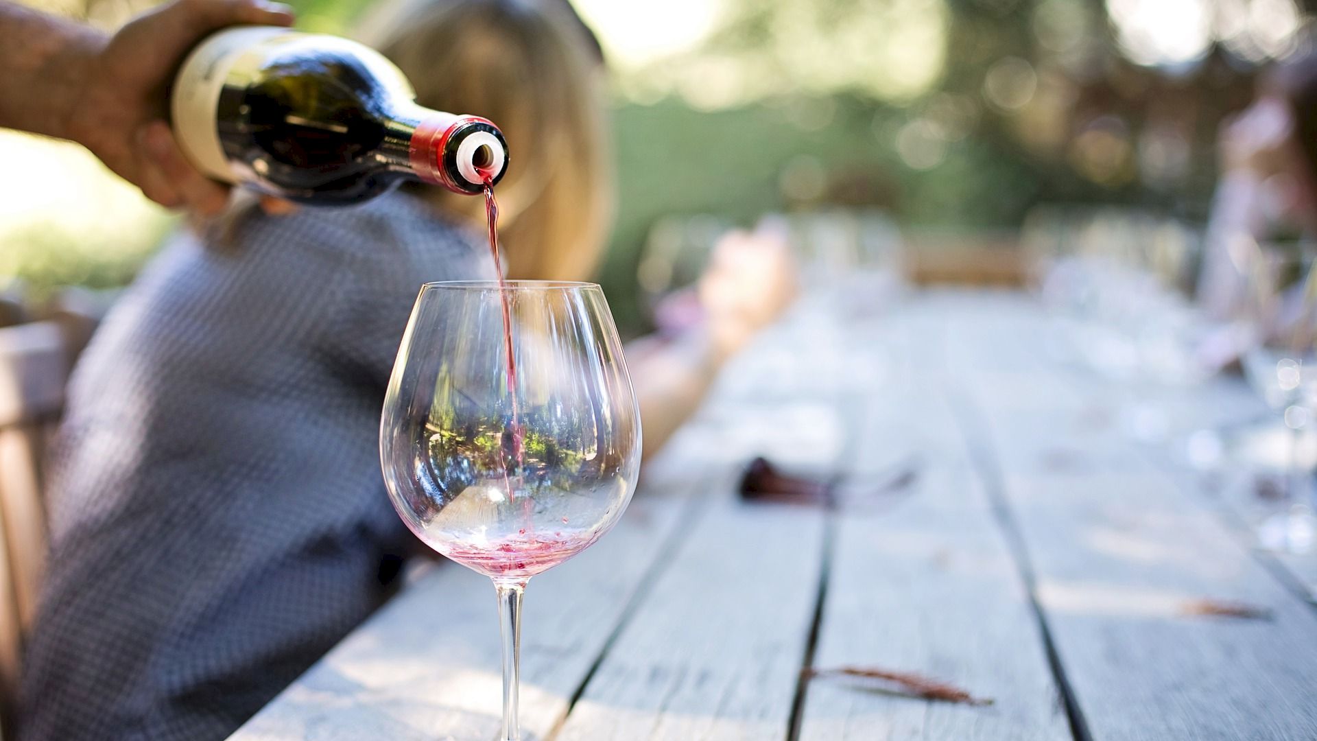 A hand pours red wine from a bottle into a wine glass on an outdoor patio table.