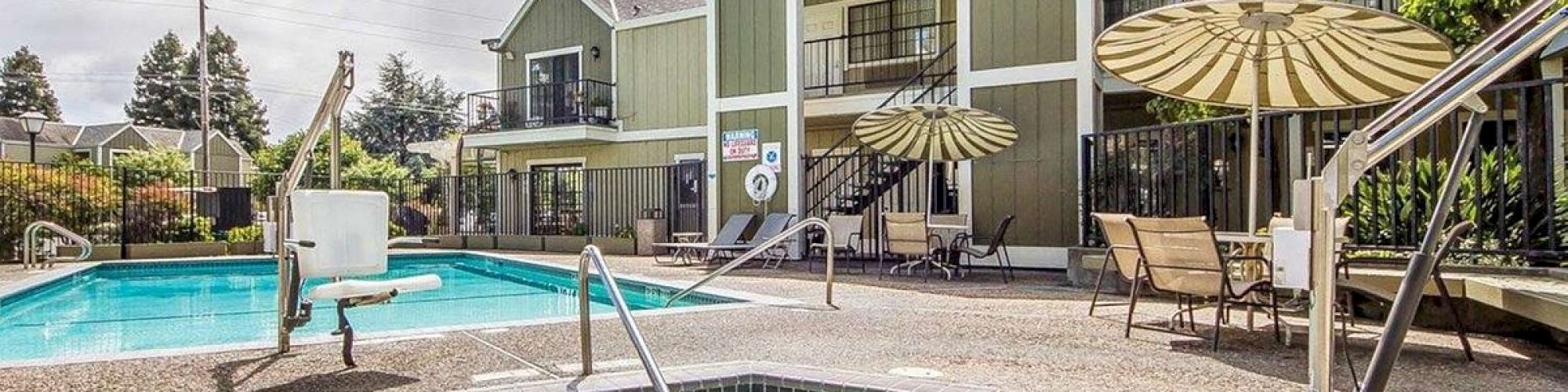 An outdoor apartment complex pool area with a hot tub, patio chairs, umbrellas, and surrounding buildings under cloudy skies.