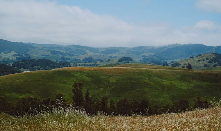 Rolling green hills and distant mountains stretch under a blue sky with scattered clouds, viewed from a grassy hillside.