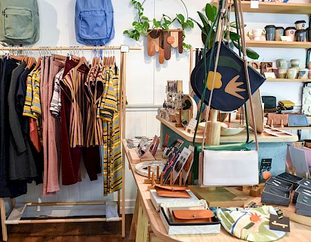 A boutique display with hanging clothes, bags, and shelves of accessories and pottery for sale.