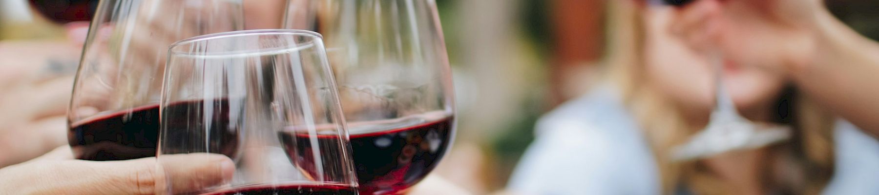 A group clinks red wine glasses outdoors, toasting together on a sunny day with friends.