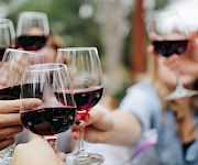 A group clinks red wine glasses outdoors, toasting together on a sunny day with friends.