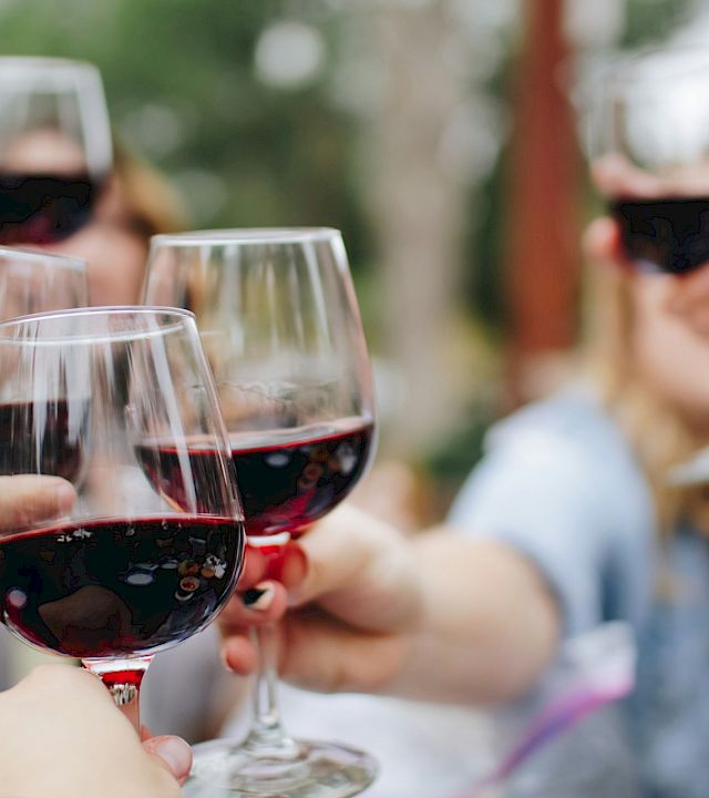A group clinks red wine glasses outdoors, toasting together on a sunny day with friends.
