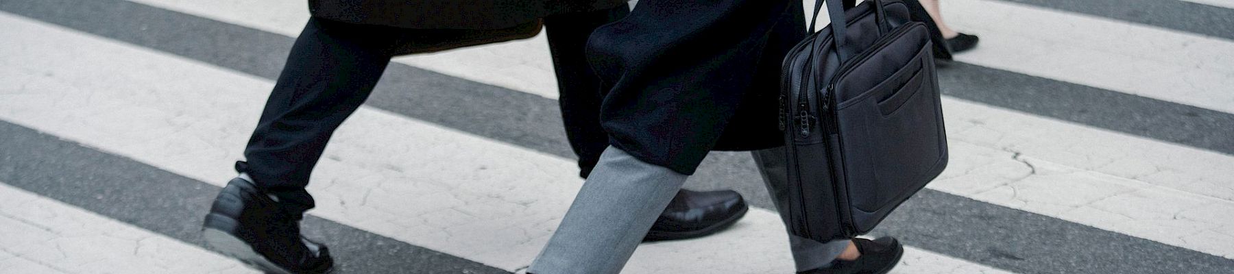 Two people in suits stride across a city crosswalk, one carrying a black briefcase and another a bag.