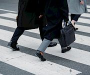 Two people in suits stride across a city crosswalk, one carrying a black briefcase and another a bag.