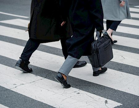 Two people in suits stride across a city crosswalk, one carrying a black briefcase and another a bag.
