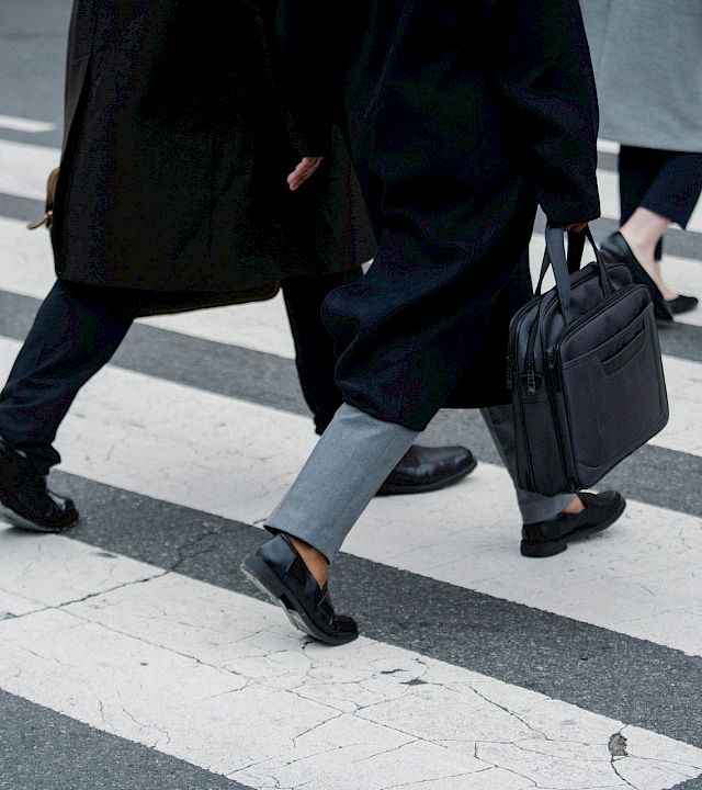 Two people in suits stride across a city crosswalk, one carrying a black briefcase and another a bag.