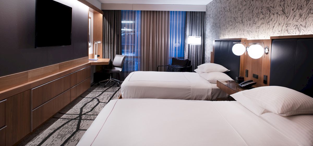A modern hotel room with two white beds, a desk, and a TV, lit by wall lamps and curtains by a large window.