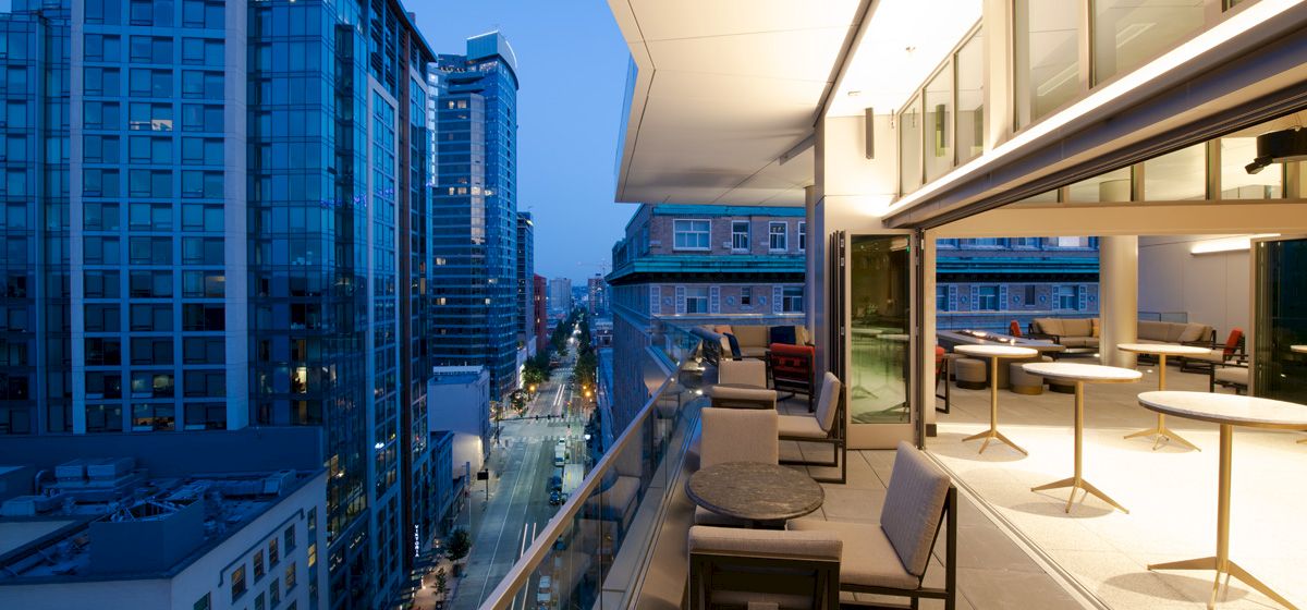Modern high-rise balcony lounge overlooks a downtown street and skyline at dusk, with chairs and caf&eacute;-style tables.